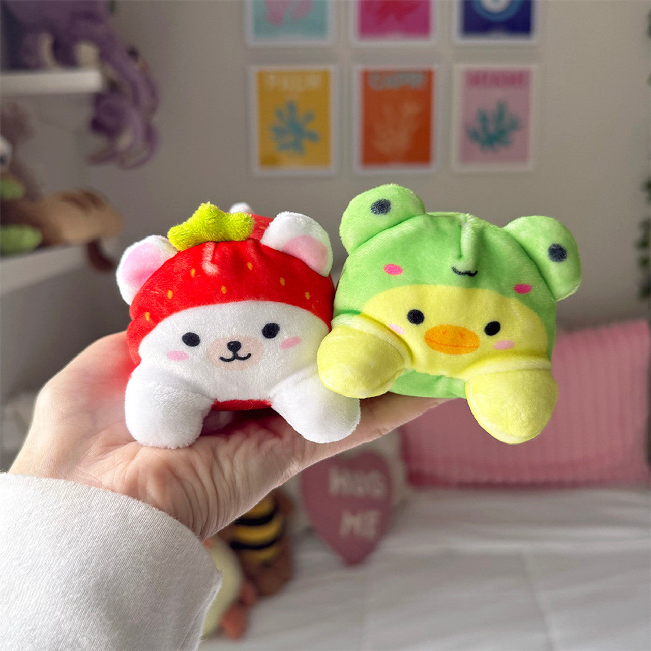 Person holding two Sand-A-Roos Sensory Plush toys, a strawberry bear and a green frog. Soft and playful designs.