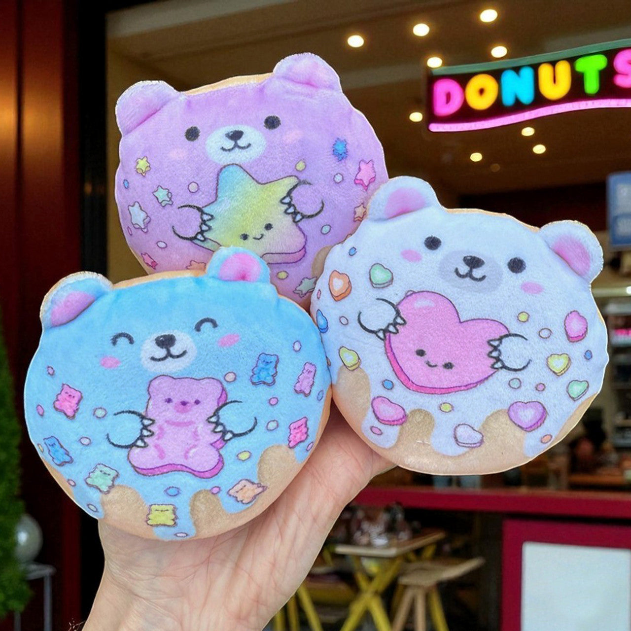 Super Duper Sugar Squishers - Plush Sugar Bear Donuts Edition by Top Trenz - Hand holding three adorable Super Duper Sugar Squisheen plush toys shaped like pastel bears with cute candy and star designs, displayed in front of a colorful donut shop.