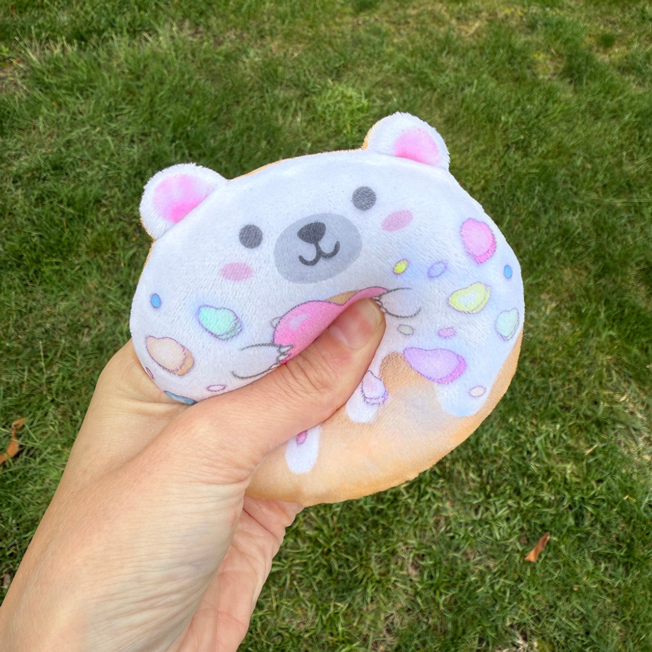 Super Duper Sugar Squishers - Plush Sugar Bear Donuts Edition by Top Trenz - Person squeezing a white bear-shaped Super Duper Sugar Squisheen plush toy decorated with pastel hearts and sprinkles on a grassy background.